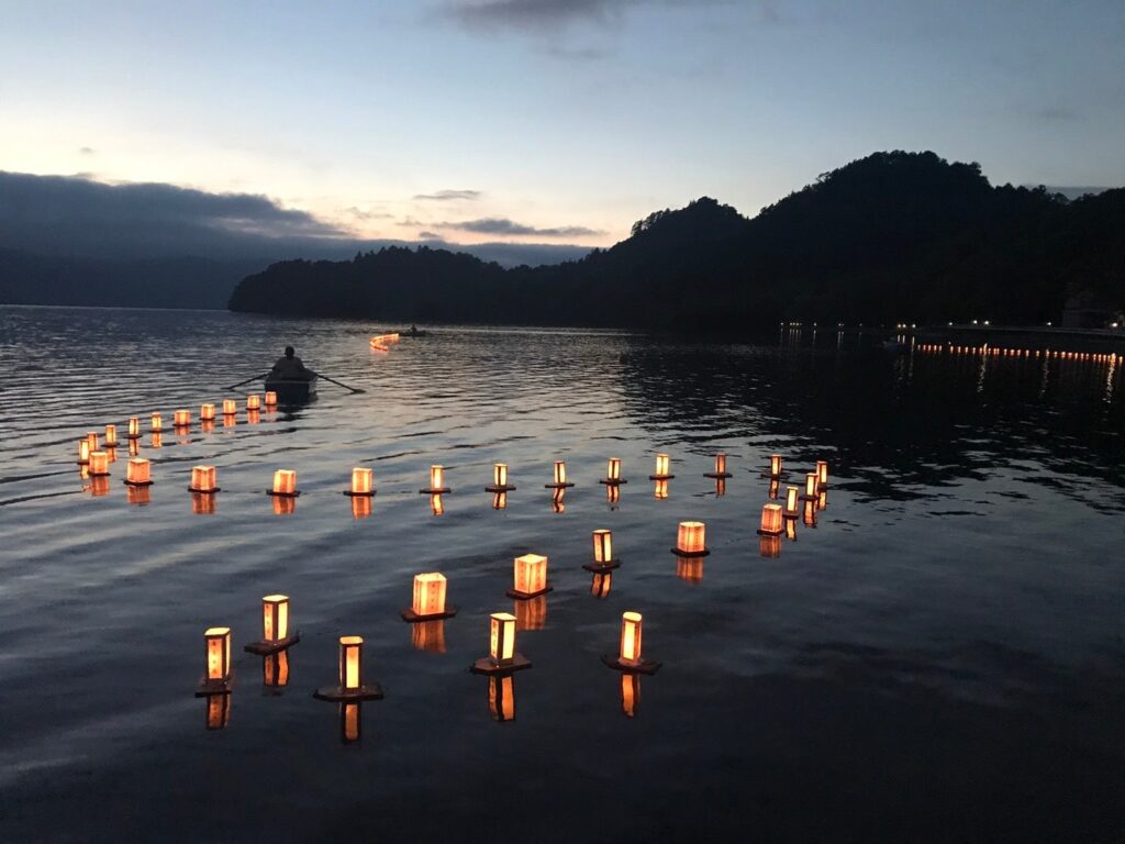 ※写真は、十和田湖畔休屋地区で毎年お盆明けに行われている灯ろう流しです。湖水まつり当日は、手漕ぎボートで願いが書き込まれた灯ろうをけん引します。